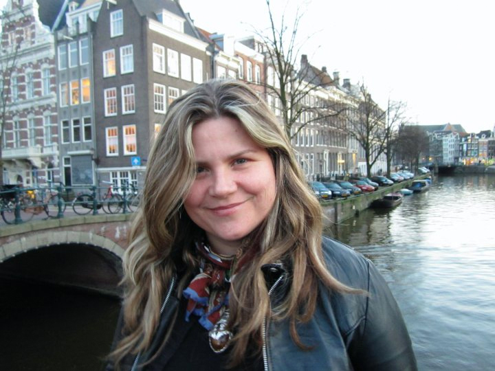 Photograph of a caucasian woman with light brown hair looking at camera, smiling. Tall historic buildings on a canal in Amsterdam are visible behind her.